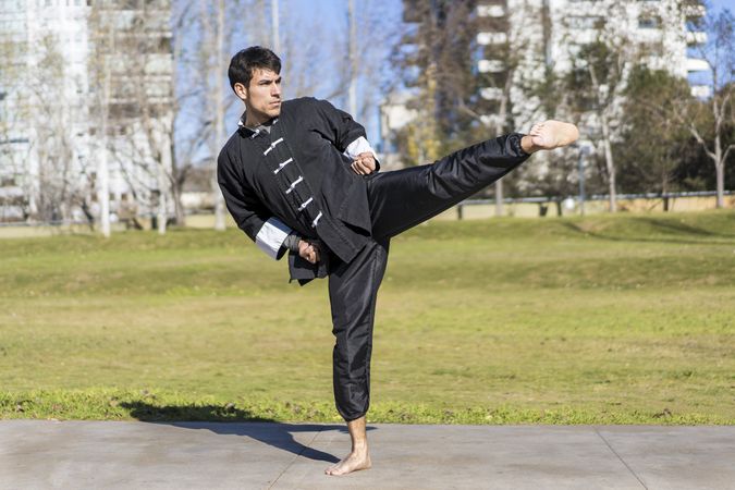 Young athletic martial arts fighter practicing kicks in a public park wearing classic kung fu uniform