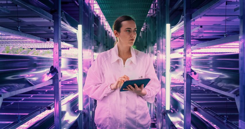 Portrait of Female Agricultural Engineer Walking in Vertical Farm Facility Next to Racks with Fresh Natural Crops. Professional Hydroponics Specialist Using a Tablet, Studying, Cultivating Plants.