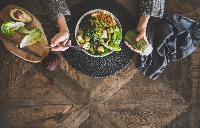 Healthy vegetarian bowl pictured on table with smoothie on side and hands with fork, copy space