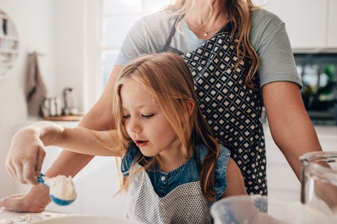 Young girl scooping flour into bowl with mother watching