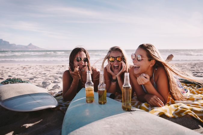 Group of young women sitting in the sand with a surfboard and beer