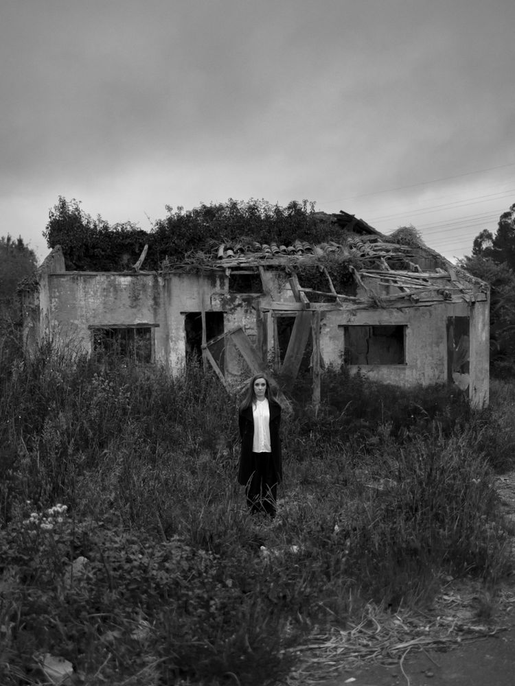 A young woman stands alone in a rugged setting, surrounded by tall grasses and an abandoned, crumbling building in the background