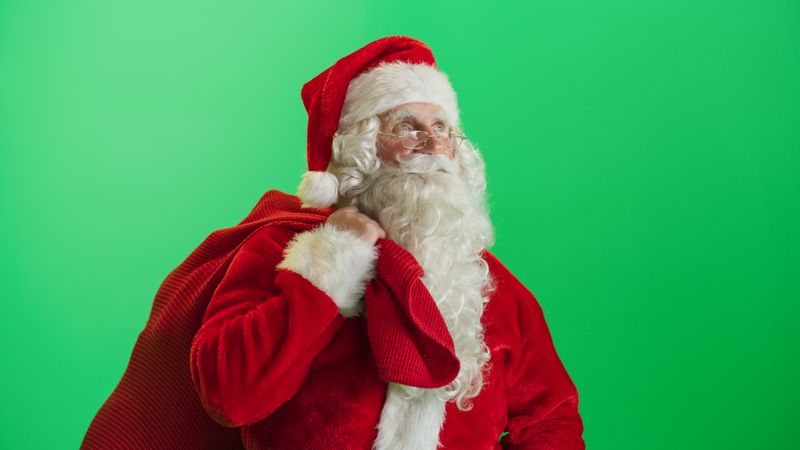 Studio Shot Isolated Mockup Template: Jolly Santa Claus Standing On Green Screen Chromakey Background And Holding Bag With Gifts, Looking at Camera. Christmas, New Year, Holiday Celebration