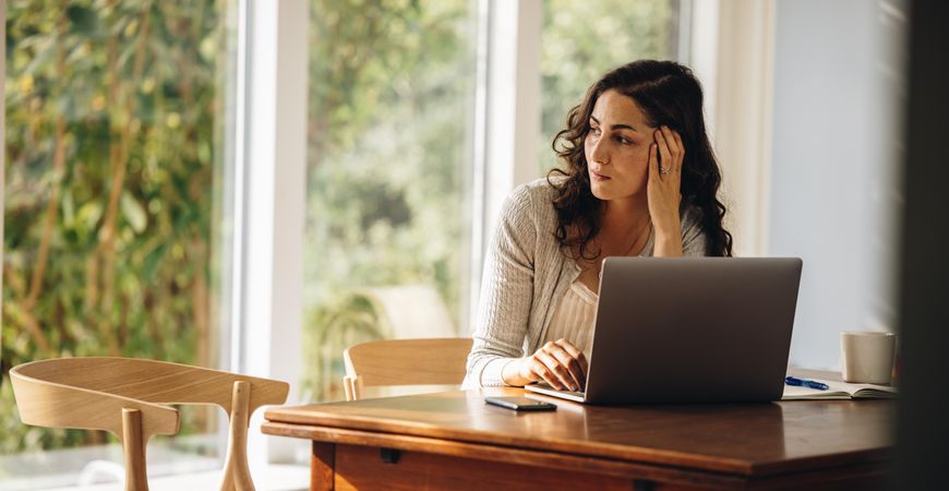 Woman sitting at kitchen table with laptop and concerned expression