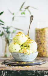 Bowl of melting pistachio ice cream with spoon, and light background with leaves and glass jar