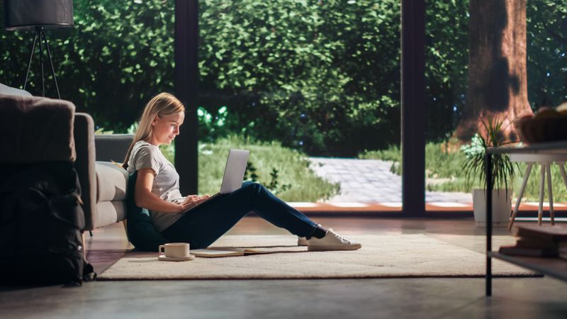 Portrait of Beautiful Young Adult Female with Blond Hair Sitting on a Floor and Using Laptop Computer in Sunny Living Room.