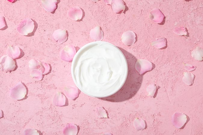 Jar with cream and rose petals on pink background, top view