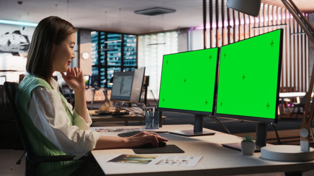 Female Asian Game Designer Using Desktop Computer With Green Screen Chromakey on Display, Designing Characters In 3D Modeling Software For Survival Video Game.