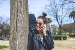 Side view of a young laughing hipster woman standing and leaning on a tree at park in sunny day while using a mobile phone
