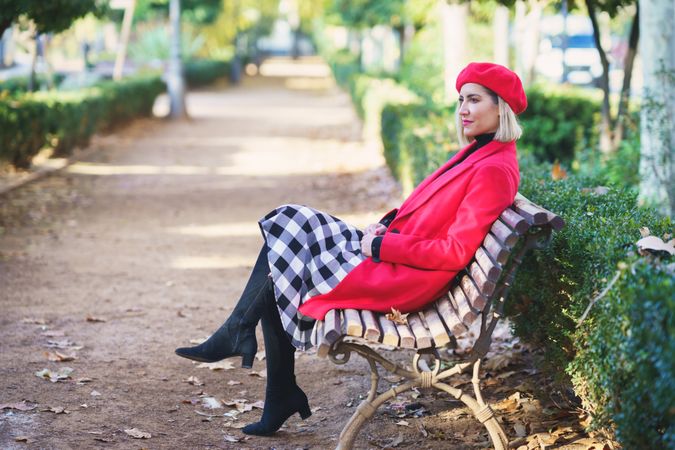 Female in red outfit and beret sitting on wooden bench in autumn park with green plants