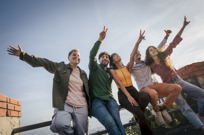 Group of diverse young friends celebrating with arms up against a blue sky