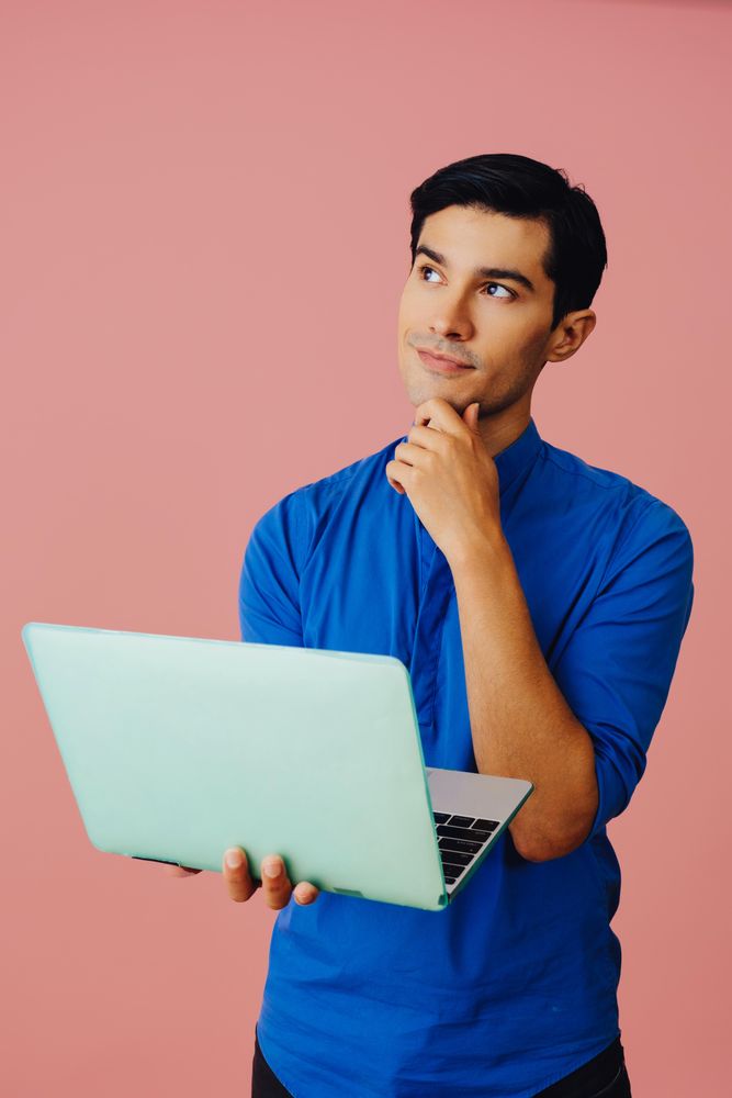 Vertical shot of contemplative Latino man looking up while holding laptop in pink room, copy space