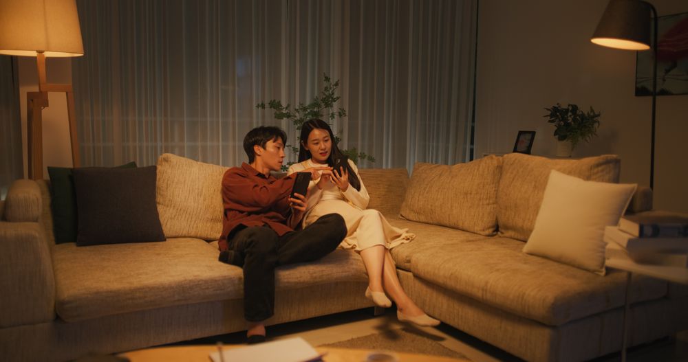 South Korean Couple Enjoying Leisure Time at Home in the Evening, Both Using Smartphones For Online Shopping