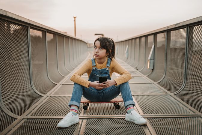 Girl with smartphone sitting on skateboard on walkway