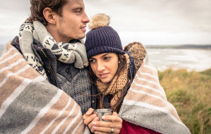 Couple cuddling while wrapped in blanket on a windy day on a cliff above the coast