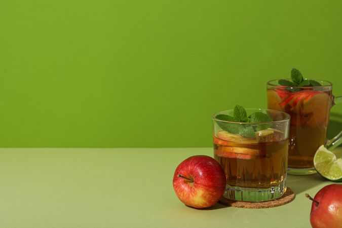 Glasses with apple cider, red apples and piece of lime on green background, space for text