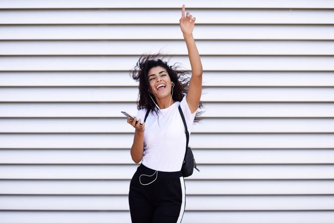 Arab woman in sport clothes with curly hair standing in front of wall dancing while pointing up