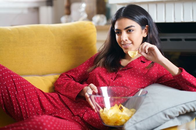 Female in red pajamas relaxing on sofa and eating chips