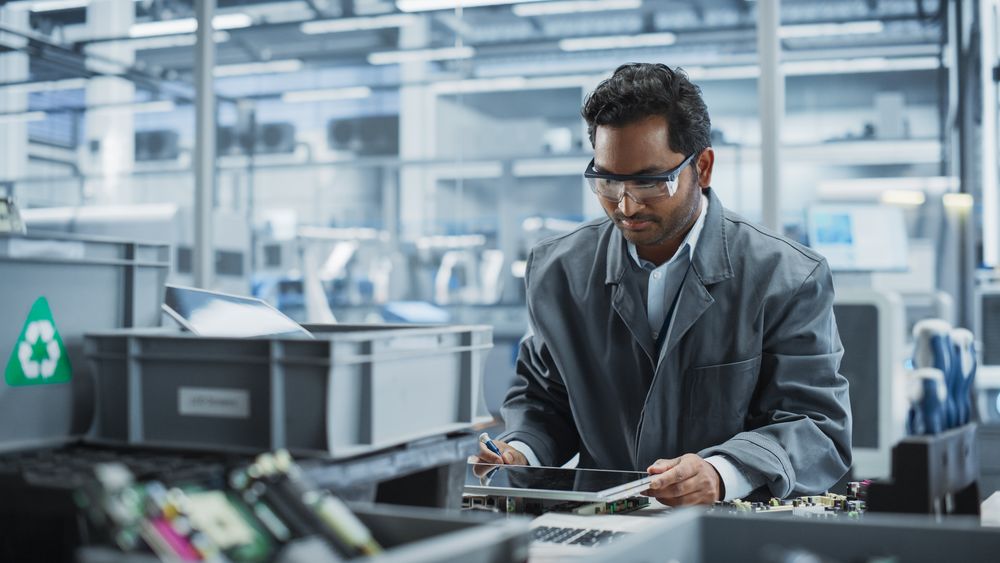 Indian Male Worker Disassembling Laptops To Recycle Electronic Components For Microchip Production At an Automated Electronics Factory With Robotic Arms.