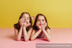 Twin girls lying on pink floor with hands on chin and looking at camera over yellow background 4BRmB4