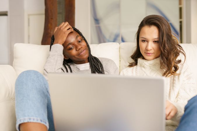 Two women in jeans watching something together on a sofa on a pc