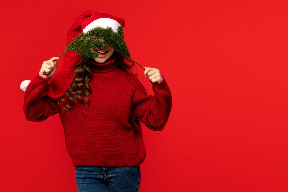 Girl in Santa hat playfully covering face with fir branches.
