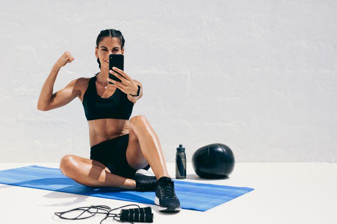 Woman in fitness wear sitting on yoga mat taking a selfie showing her biceps