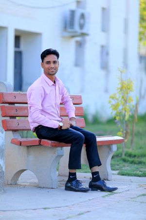 Man in pink dress shirt and dark pants sitting on a bench outdoor