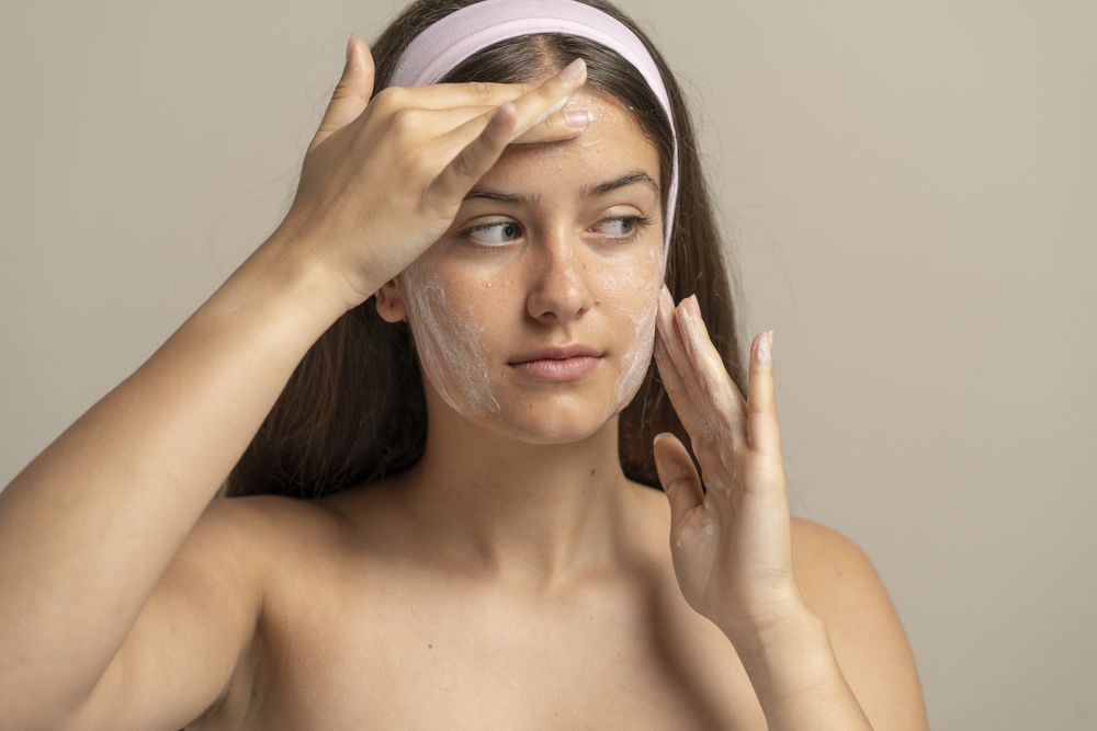 Young woman in studio gently applies white foam cleanser mask to her bare face, showcasing natural skin texture and visible pores in a realistic daily skincare ritual.