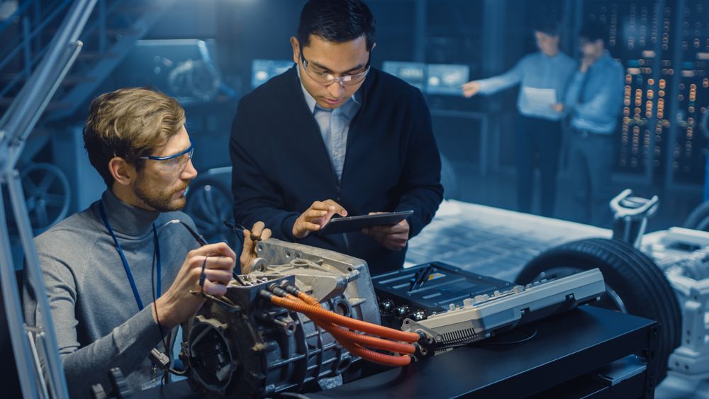 Two Professional Automotive Engineers with a Tablet Computer and Inspection Tools are Having a Conversation While Testing an Electric Engine in a High Tech Laboratory with a Concept Car Chassis.