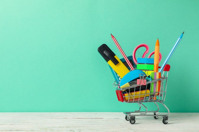 Shopping trolley with school supplies on mint background, space for text