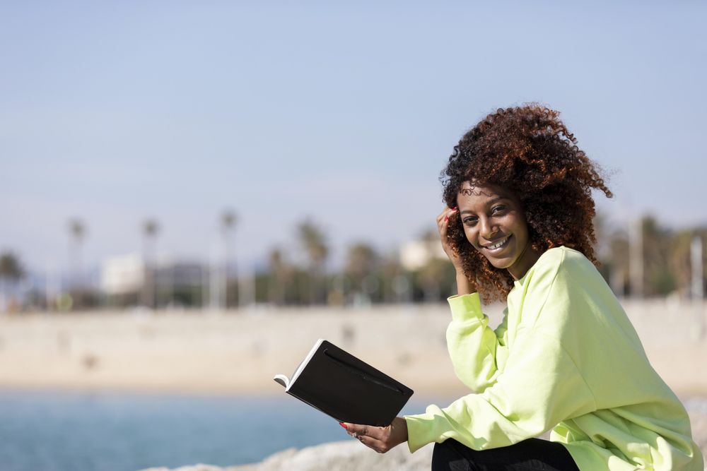 Happy female in bright green shirt enjoying reading a book sitting by the ocean