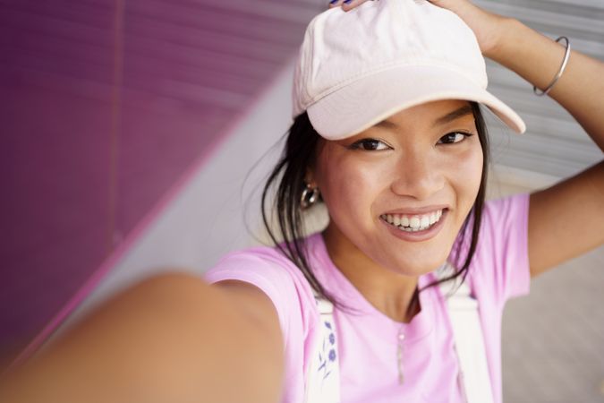 Laughing Chinese female looking up at camera while taking a selfie of herself next to pink wall