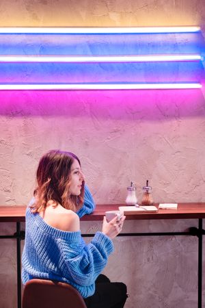 Positive woman with cup at table near wall with neon lamps
