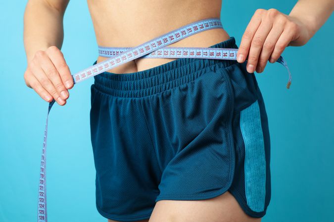 Woman in sportswear measuring her waist on blue background. Healthy lifestyle