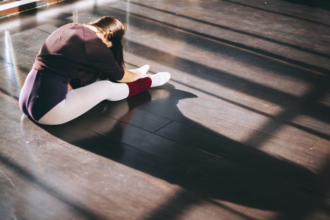 Back view of woman sitting on sunny floor of studio and warming up body before dance.