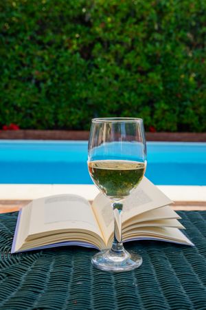 A glass of wine on an open book by the swimming pool, concept of a luxury summer vacation