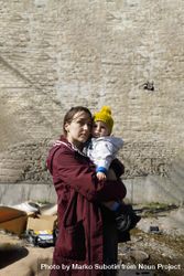 Woman holding her baby looking at rubble in front of brick wall 4NL9e0