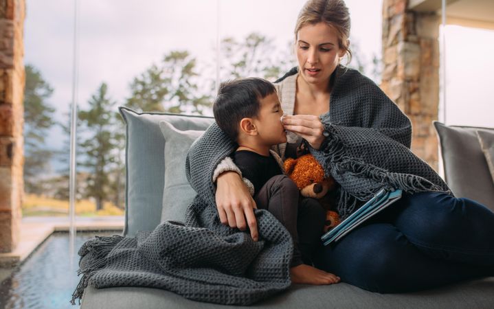 Mother wiping nose of her little son while wrapped in blanket on the sofa