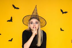 A woman in a Halloween costume shows "shh", a silent gesture, on a yellow background