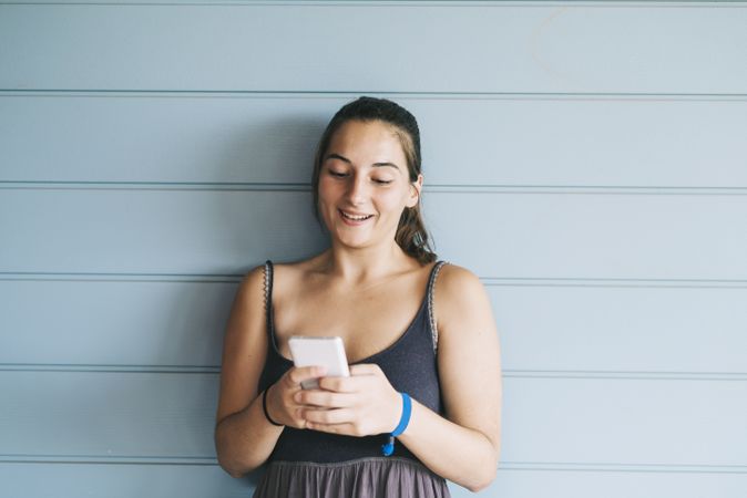 Happy teenage female using cell phone while leaning against wood paneled wall