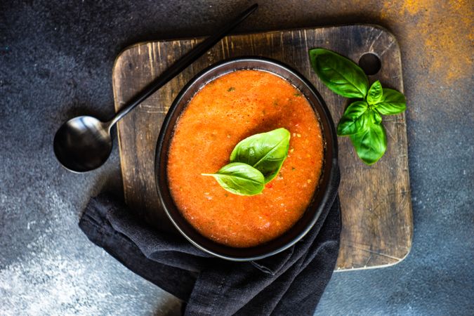 Top view of dark bowl of gazpacho soup with basil leaves and wooden board