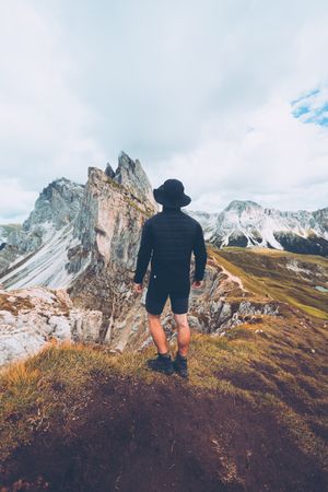 Back view of hiker wearing dark hat standing facing mountainous landform in Italy