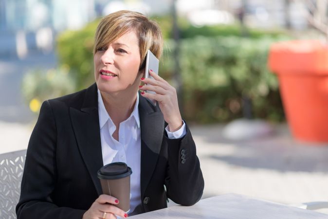 Woman sitting on table outside with coffee speaking on cell phone