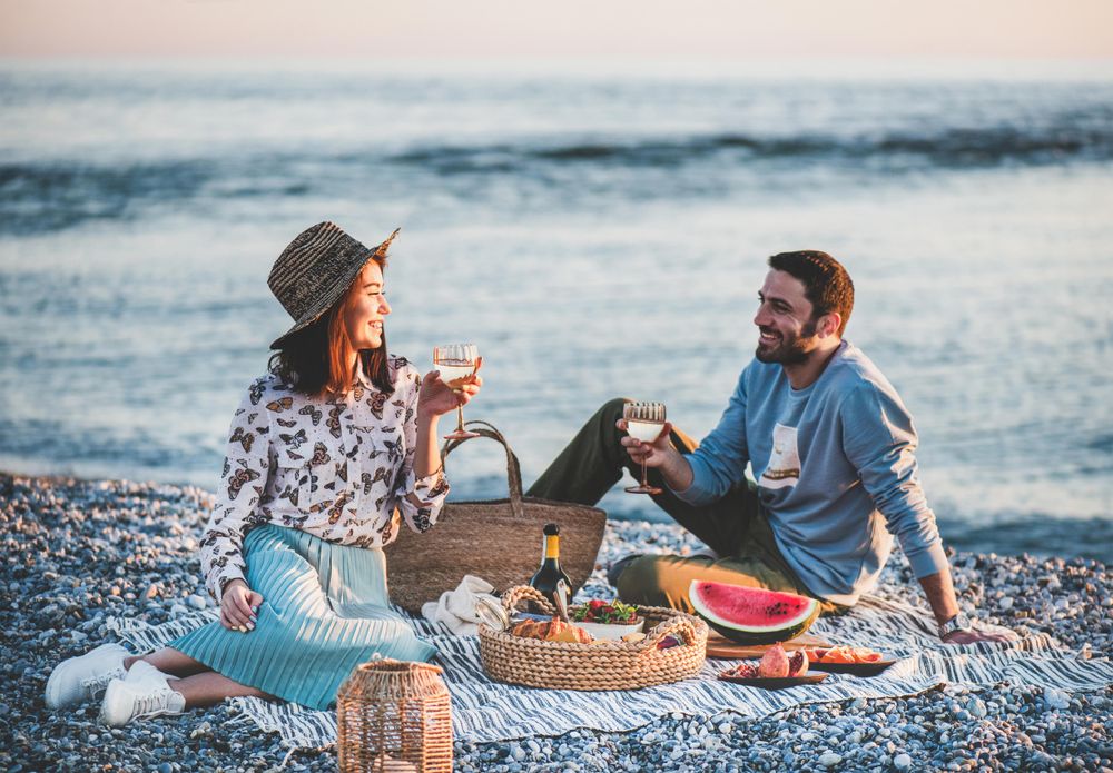 Couple smiling holding wine glasses over summer picnic by the ocean