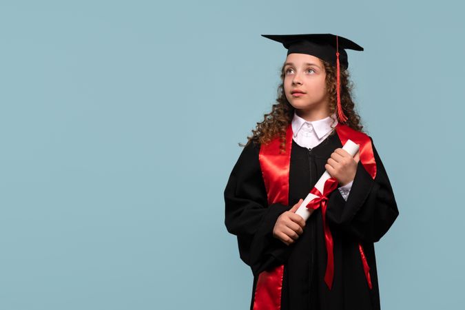 Girl in graduation gown looking up thoughtfully while holding diploma.
