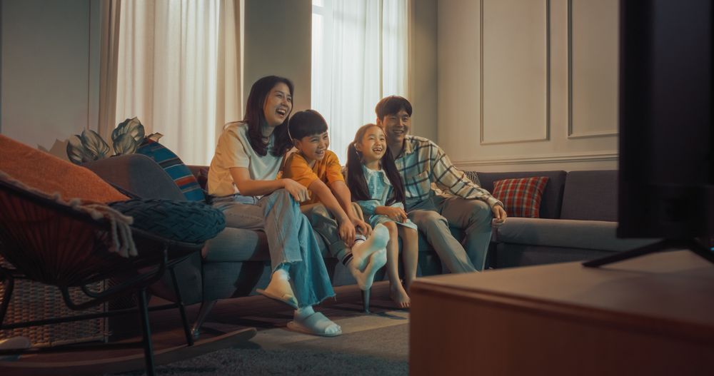 Happy Korean Family At Home: Mother And Father Watching Funny Family Television Show And Laughing With Their Little Son And Daughter On The Couch.