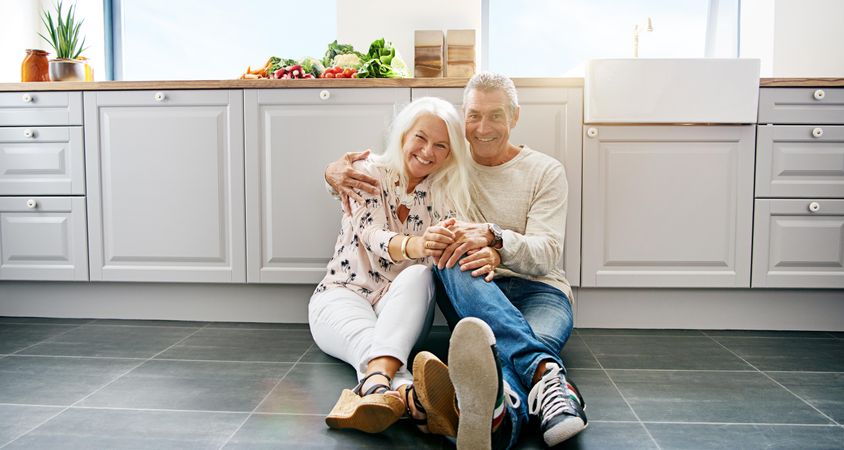 Smiling grey haired couple sitting on a bright kitchen floor
