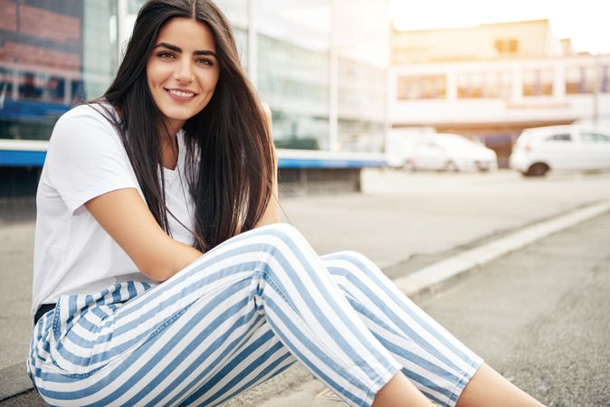 Smiling woman sitting on curb outside in striped blue pants