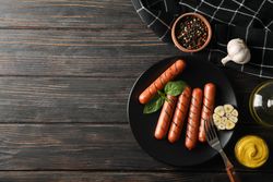 Top view of grilled sausages or hot dogs on a fork on dark ceramic plate with garlic, copy space
